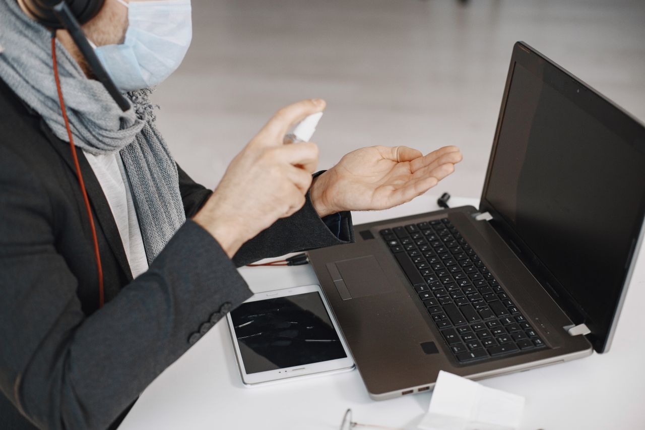 A man wearing a mask disinfecting his hands while working on a laptop indoors.