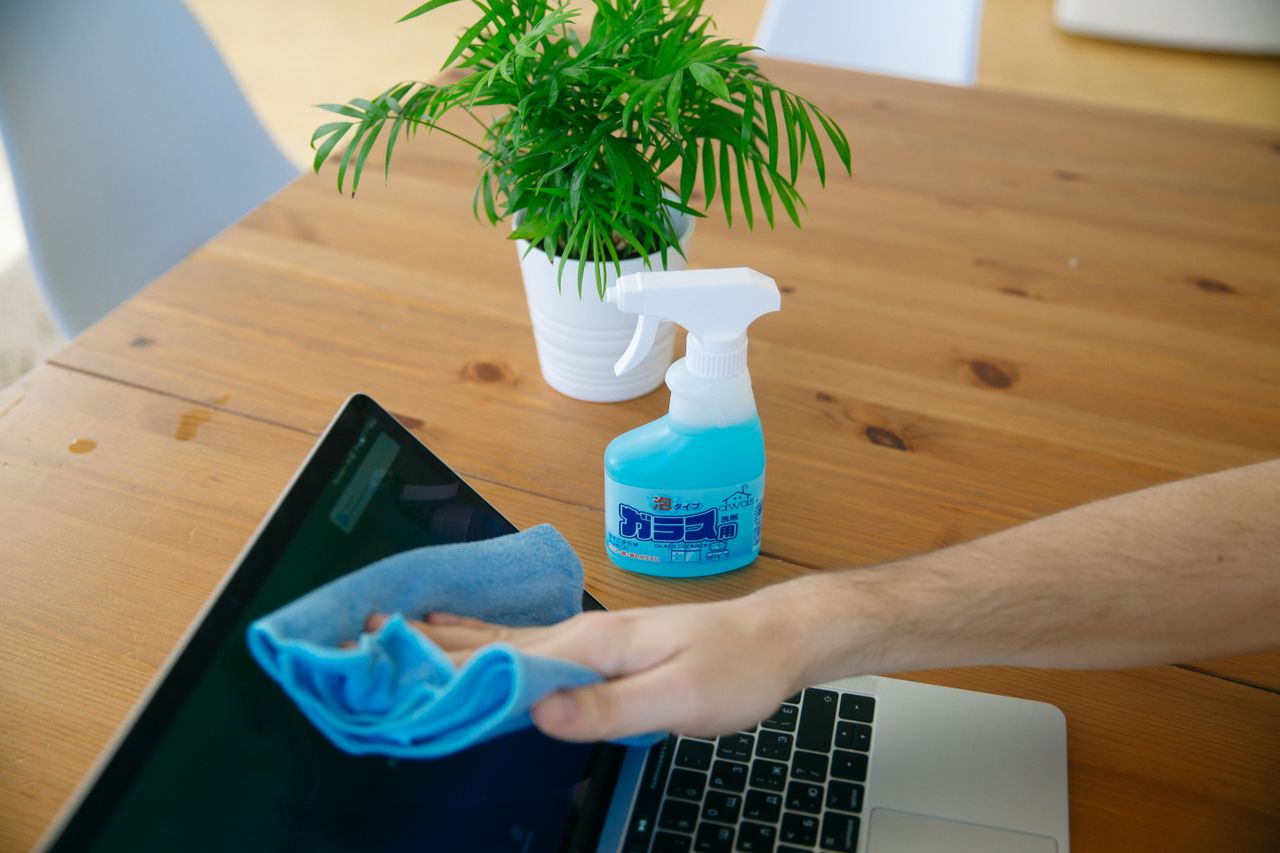 Vue rapprochée d’une main nettoyant l’écran d’un ordinateur portable avec un chiffon bleu et un flacon pulvérisateur sur un bureau en bois.